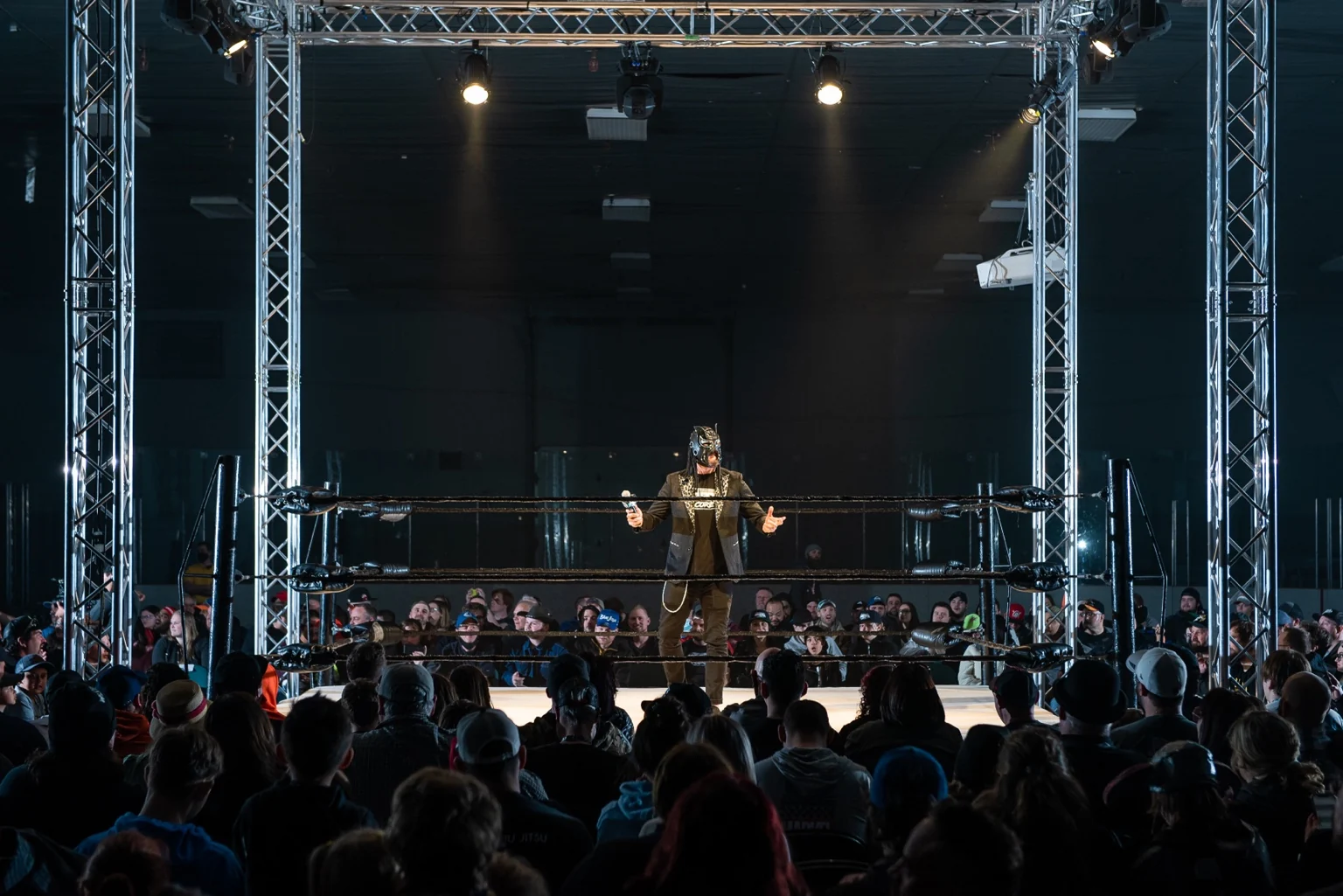A masked host addresses a crowd surrounding the WrestleCore ring.