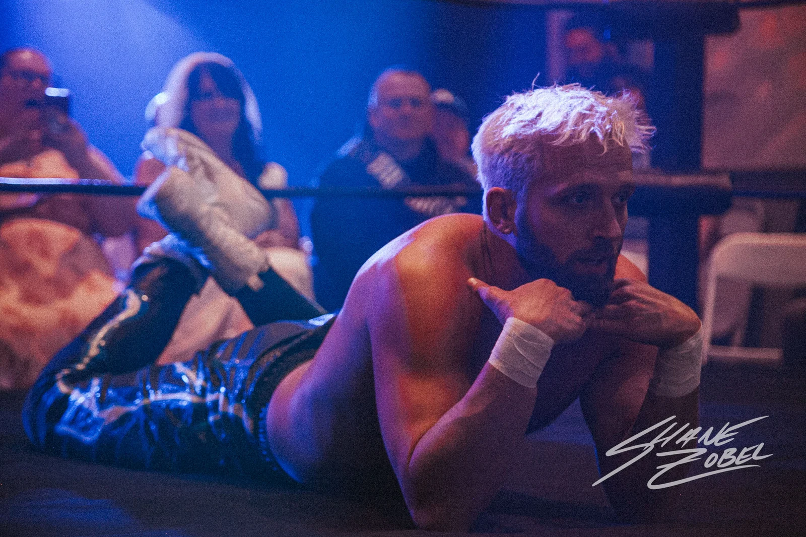 A wrestler reacts near the ropes with fans watching from ringside.